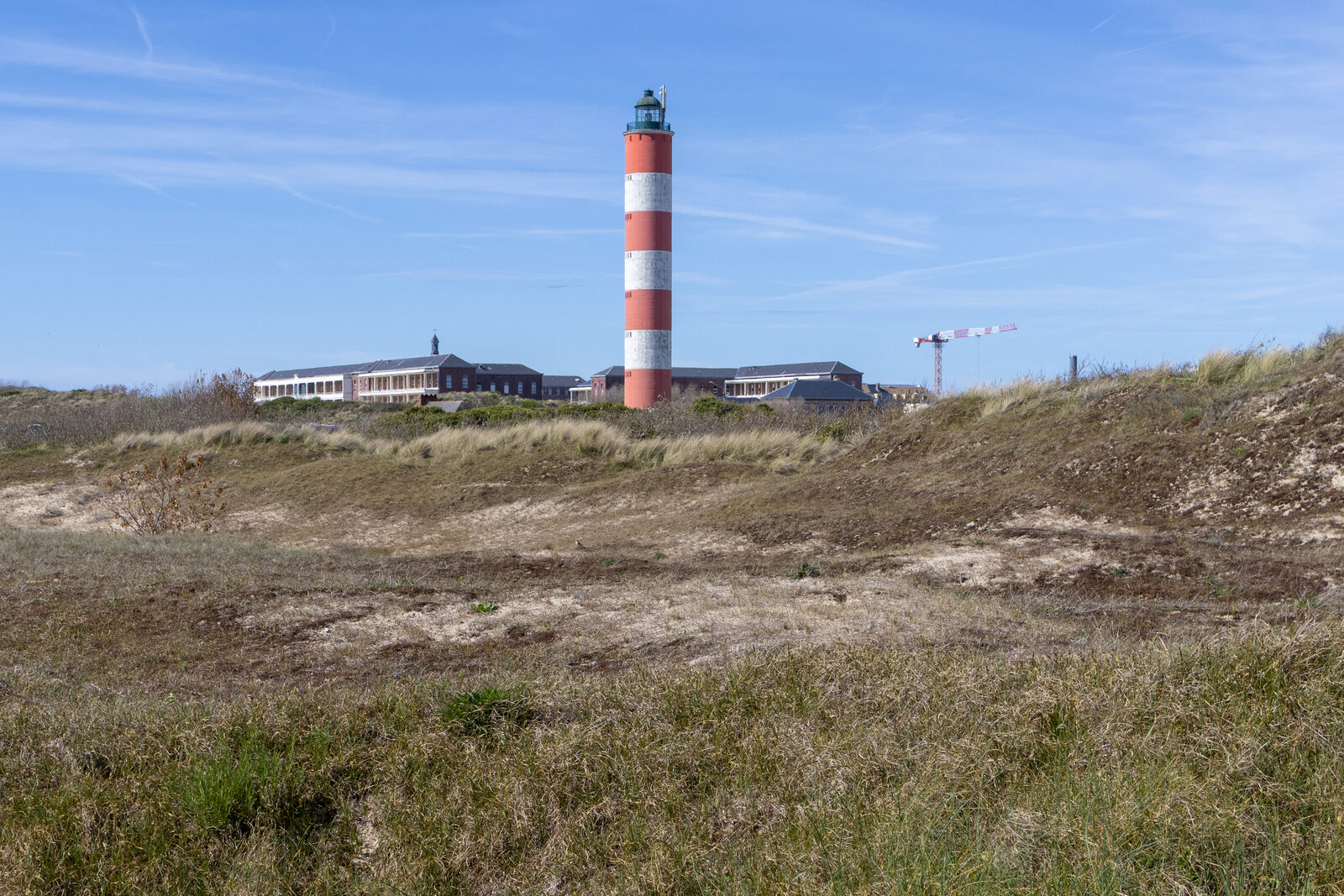 Le phare rayé rouge et blanc de Berck, vu depuis les dunes sous un ciel bleu, avec des bâtiments en brique à l’arrière-plan