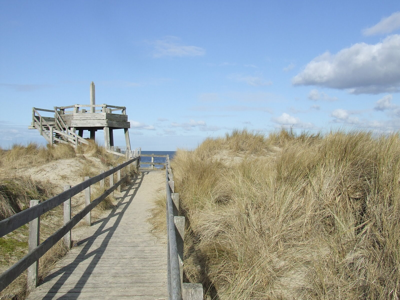 Un chemin en planches de bois avec garde-corps traverse les dunes et les oyats jusqu’à un belvédère en bois, face à la mer, côté Le Touquet de l’estuaire de la Canche